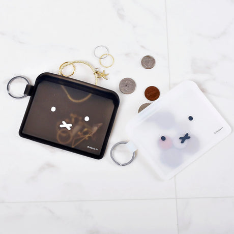 Two small pouches, one black and one semi-transparent white resembling a Miffy Bunny Card Case, feature simple faces. The black pouch holds jewelry, while the white silicone pouch seems empty. Nearby, coins and rings are scattered on a marble surface.