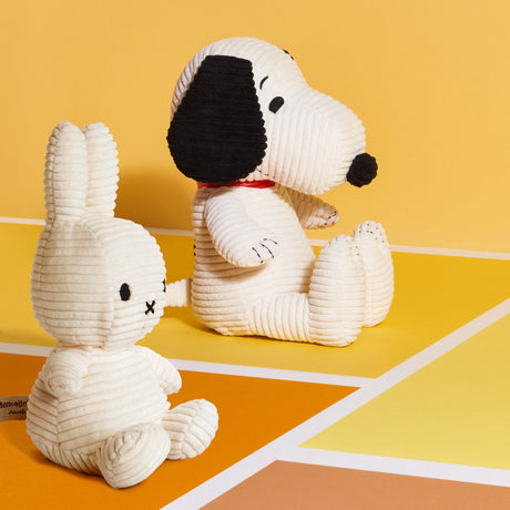 Two soft, baby-safe plush toys—a Miffy Bunny Plush Corduroy in white and a white dog with black ears—sit on a yellow and orange checkered surface against a yellow background.