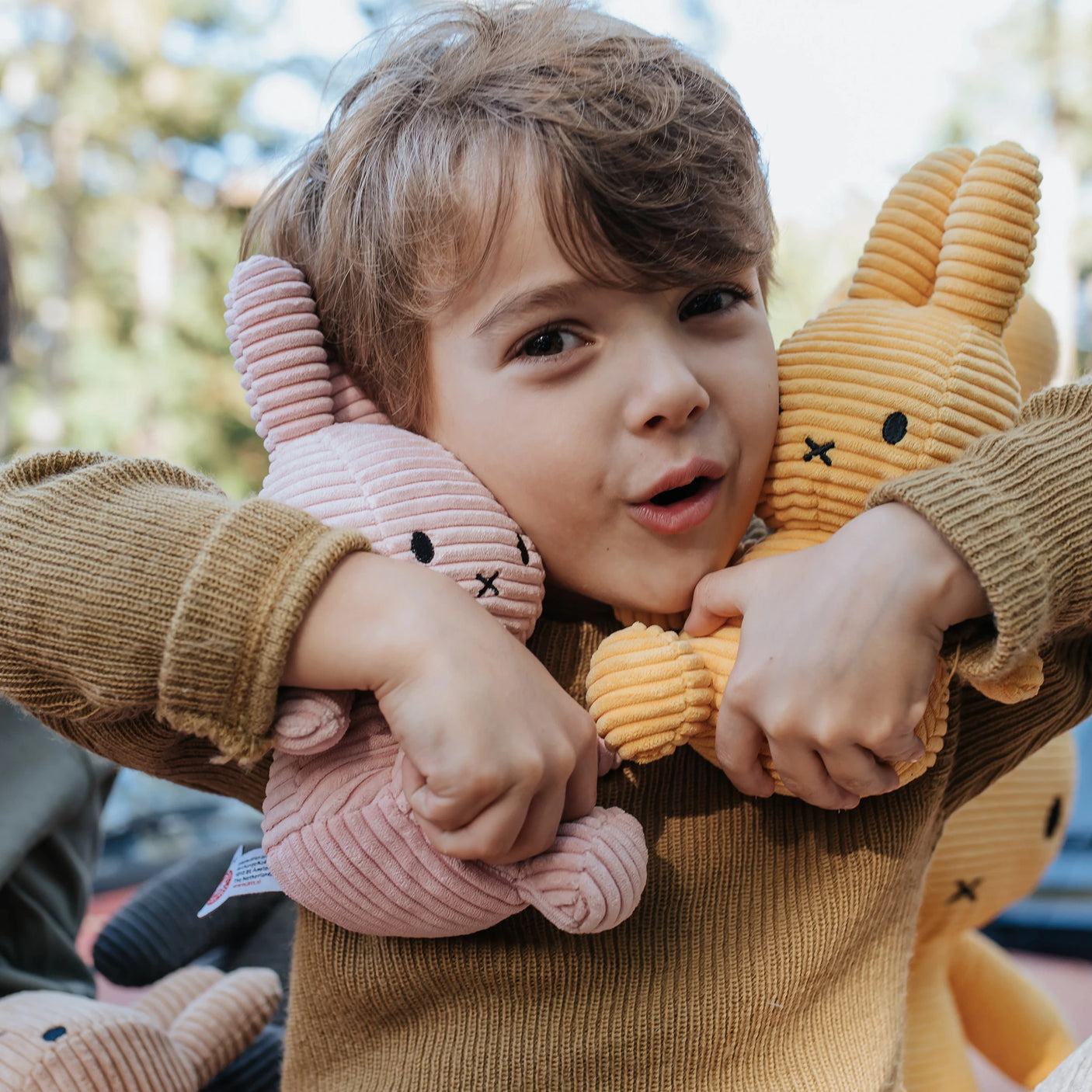 Outdoors, a child wearing a brown corduroy sweater holds two Miffy bunnies, one pink 23cm Miffy Bunny Plush Corduroy and the other yellow.