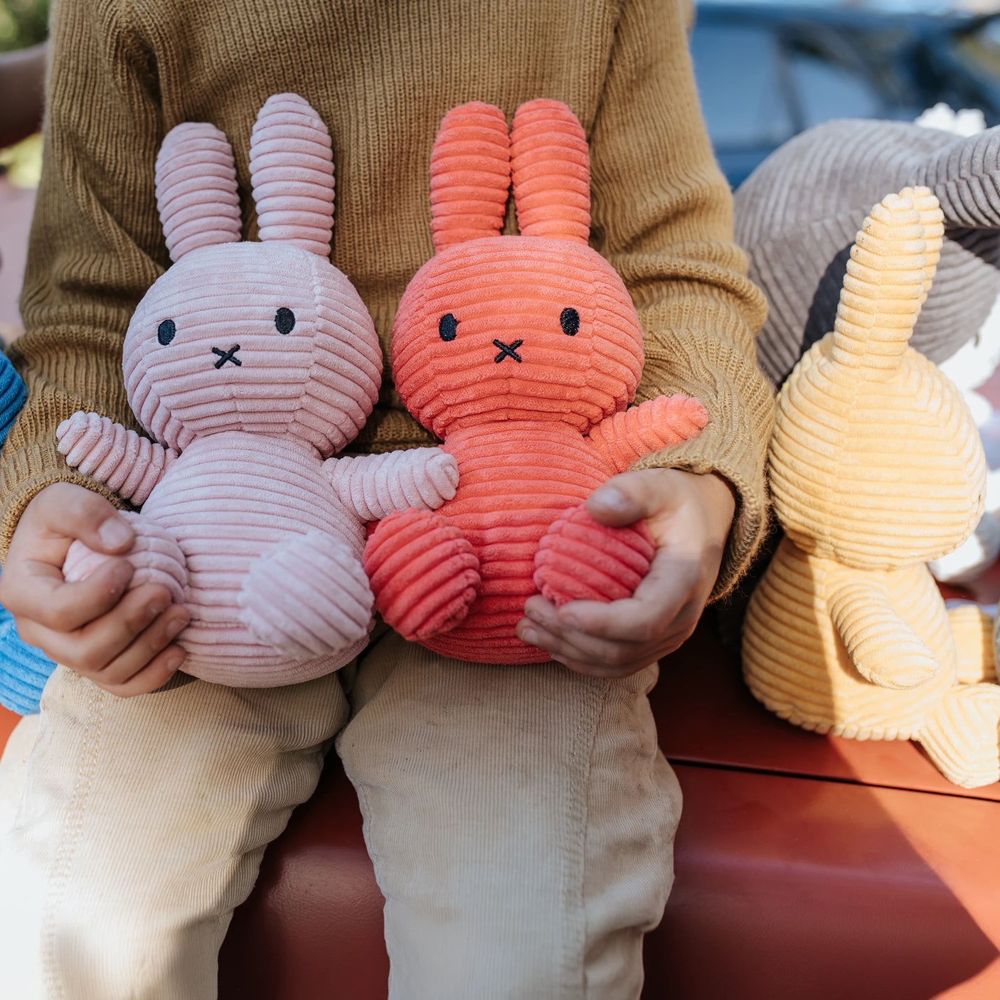 Seated with a Miffy Bunny Plush in each hand—one pink, one red—a person enjoys the company of these hypoallergenic companions. A Miffy Bunny Plush Corduroy in buttercream, crafted from recycled polyester, rests peacefully beside them.