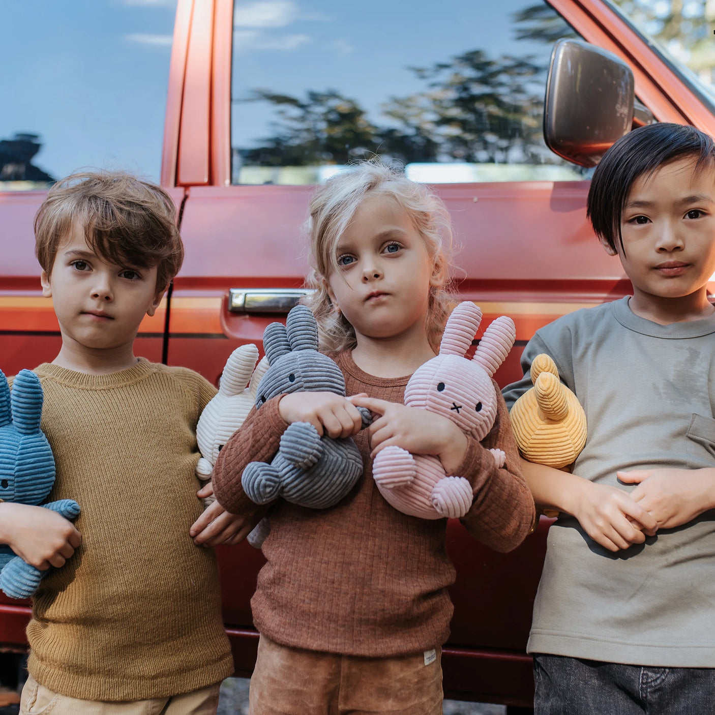 Three children holding Miffy Bunny Plush Corduroy toys in pink, measuring 23cm, stand in front of a red car.