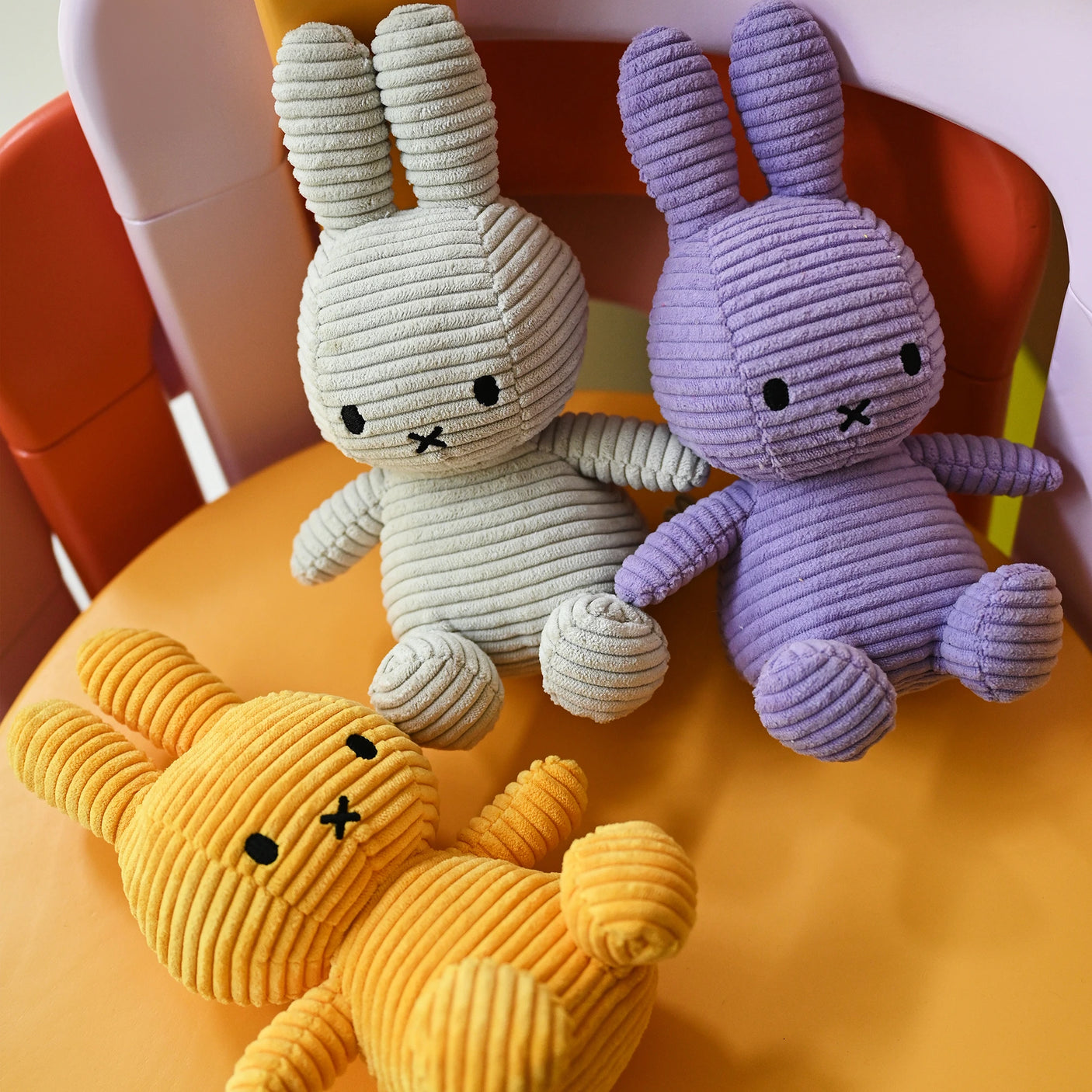 Three eco-friendly Miffy Bunny Plush Corduroy toys in gray, lilac, and yellow sit on a chair with multiple colors.