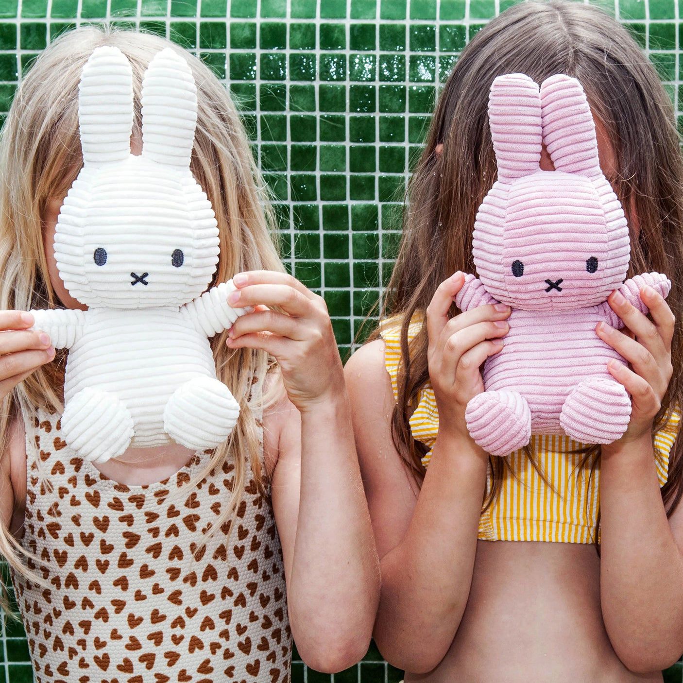 Two children holding baby-safe Miffy Bunny Plush Corduroy toys in white, 23cm, in front of their faces, standing against a green tiled wall. The child on the left is dressed in a leopard print outfit, while the one on the right is wearing yellow stripes.