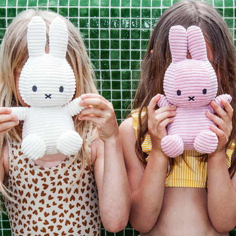 Two children holding baby-safe Miffy Bunny Plush Corduroy toys in white, 23cm, in front of their faces, standing against a green tiled wall. The child on the left is dressed in a leopard print outfit, while the one on the right is wearing yellow stripes.