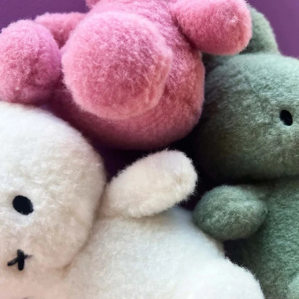 A trio of plush toy animals is shown in close-up: one white, reminiscent of a Miffy Plush, one pink that resembles the Miffy Plush Cotton Candy - Rose 23cm, and one green. They all feature simple facial expressions and are clustered together against a purple backdrop.