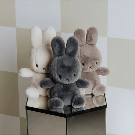 The Cozy Miffy Plush & Gift Box Cream - 23cm from the Miffy brand features three super soft plush bunny toys in white, gray, and brown. Arranged on a reflective pedestal against a checkered background, these cuddly companions are the perfect surprise for any gift box.