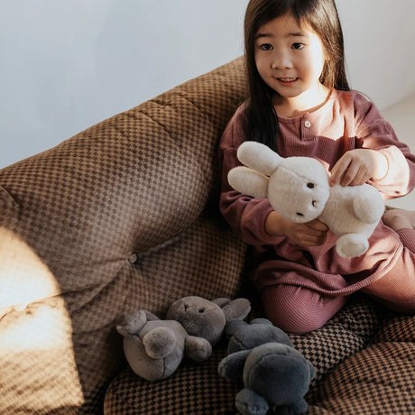 A child in a pink outfit sits on a brown patterned couch, cuddling the Cozy Miffy Plush & Gift Box Cream - 23cm by Miffy, which is crafted from super soft fabric. The white plush bunny is accompanied by three other adorable bunnies.