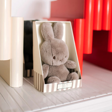 The Cozy Miffy Plush & Gift Box Taupe from Miffy, measuring 23cm and crafted from recycled polyester, is snugly presented in a striped gift box on a white shelf next to red and white cylindrical objects.