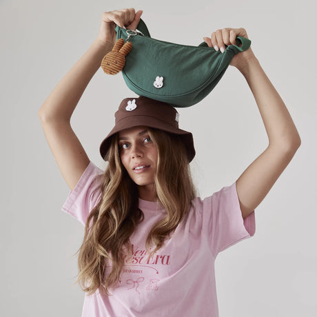 Woman holding green Miffy Crescent Crossbody Bag with plush charm above her head, wearing brown bucket hat and pink shirt, promo 1