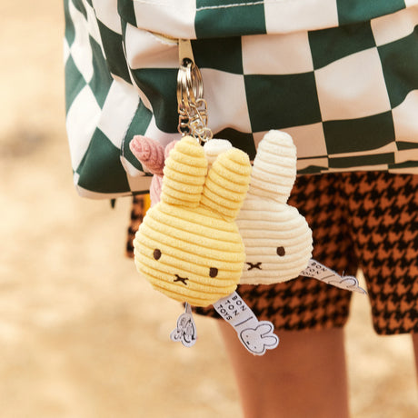 Two charming Miffy Flat Corduroy Keychains, one in off-white and the other in yellow, are attached to a checkered bag, adding an adorable touch. Made from eco-friendly materials, these Miffy-inspired bunnies perfectly complement the look with their playful and sustainable design.