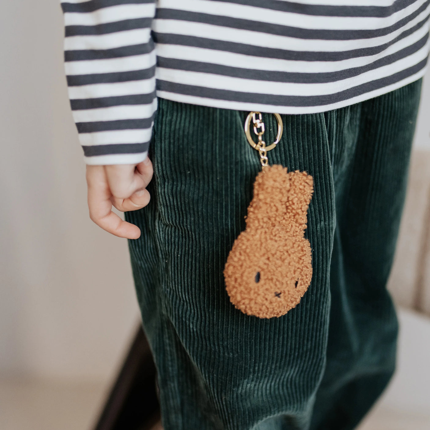 A child in a striped shirt and green corduroy pants showcases an eco-friendly flair with a Miffy Keychain Tiny Teddy Plush - Terry attached.