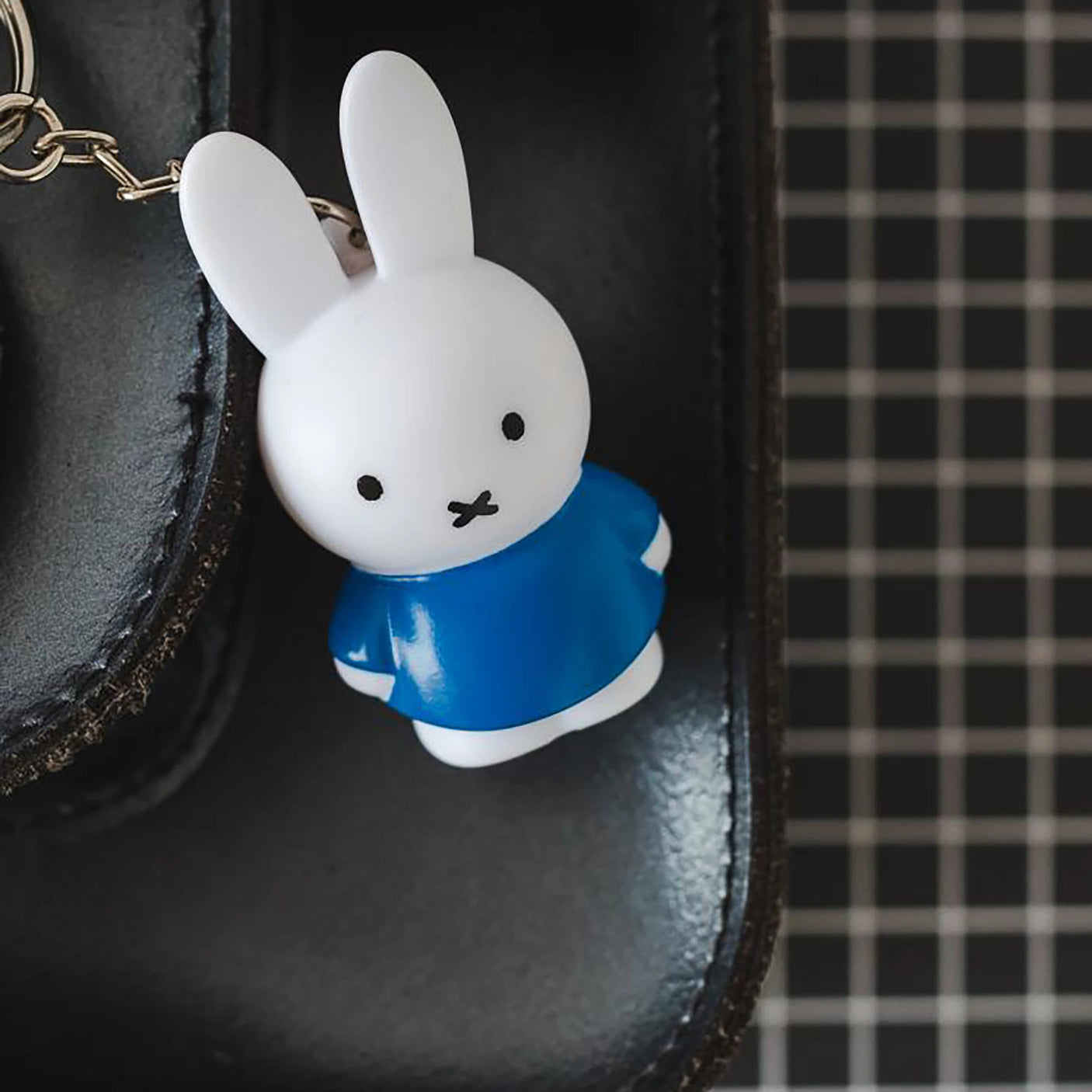 A Miffy Keychain - Classic from the Miffy brand, featuring a white rabbit in a blue dress, dangles from a black bag with a grid pattern, adding a charming touch of cute accessory flair.