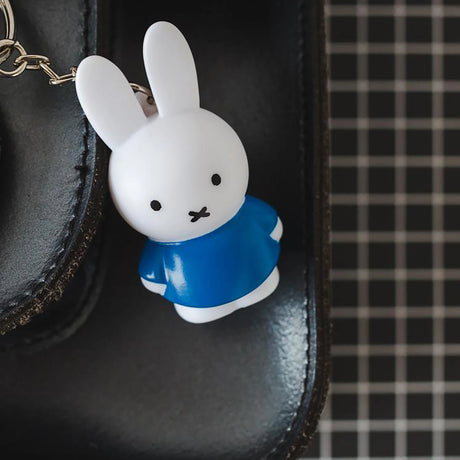 A Miffy Keychain - Classic from the Miffy brand, featuring a white rabbit in a blue dress, dangles from a black bag with a grid pattern, adding a charming touch of cute accessory flair.