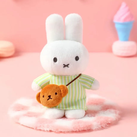 White plush bunny in green striped dress with brown bear-shaped crossbody bag on fluffy pink surface