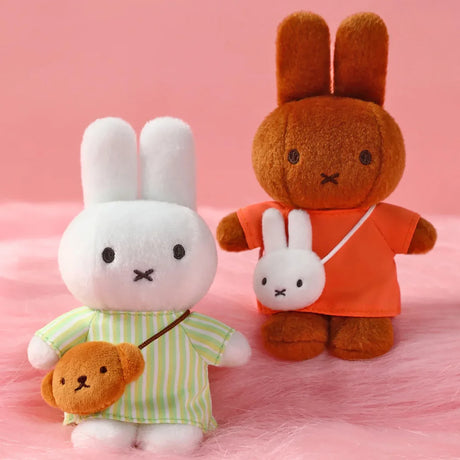 Two plush bunnies, one white in striped green shirt with brown bear bag, one brown in orange shirt with white bunny pouch on pink surface