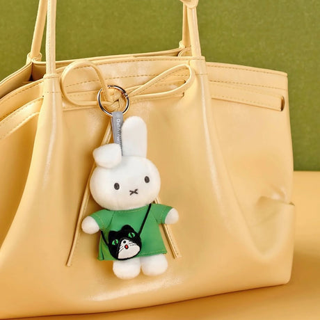 White plush bunny keychain wearing green shirt with black cat pouch hanging on beige handbag against green backdrop