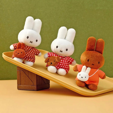 Three plush bunny toys wearing red outfits and holding small animal-shaped pouches on a beige tilted tray