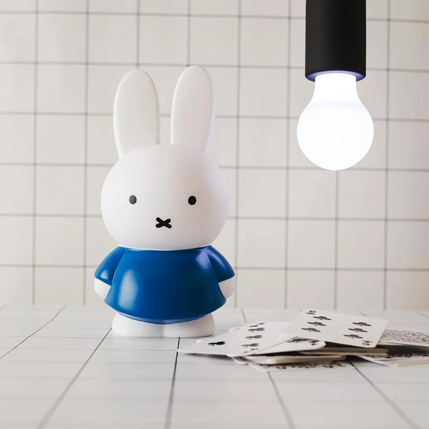 A white rabbit figurine, similar to the Miffy Bunny - Blue Money Box 12cm by Miffy, stands in a blue outfit on a tiled surface with scattered playing cards under a hanging light bulb.