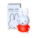 The Miffy Bunny - Red Money Box 18cm, by the brand Miffy, showcases Miffy in a delightful red dress and stands gracefully in front of its packaging, providing a stylish touch to your home decor.