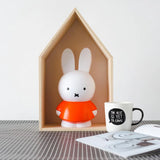 This charming Miffy Bunny - Red Money Box by Miffy serves as a stylish accent for home decor, nestled within a wooden house-shaped frame alongside a mug featuring the text "The best is yet to come" and open books on a checkered surface.