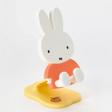 Pastel Miffy phone and tablet stand with orange shirt, white limbs, and yellow base decorated with a small fruit illustration on white background
