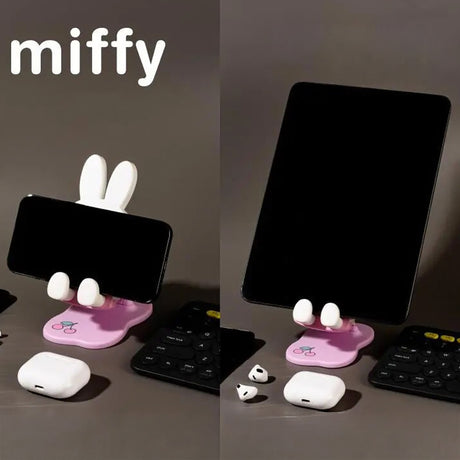 Pastel pink Miffy phone and tablet stand with white ears and hands holding devices on dark surface near wireless earbuds and keyboard