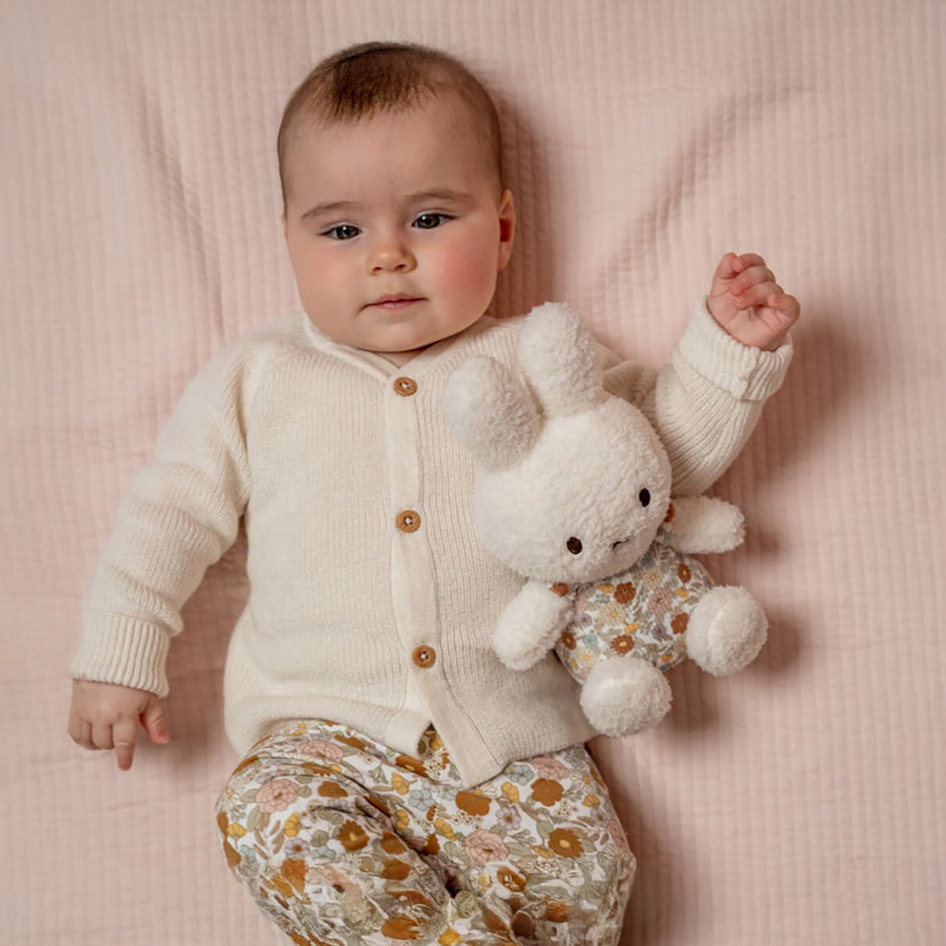 A baby wearing a white sweater and floral pants is lying on a pink blanket, cuddling the Miffy Plush White & Vintage Flowers toy by Miffy.