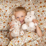 A child beams with joy, holding two Miffy 25cm plush toys from the Vintage Flowers All Over collection, nestled amidst floral-patterned bedding.