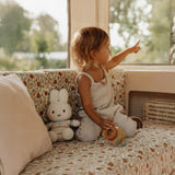 A child sits on a vintage sofa with sunny stripes, holding a toy and pointing out a window, while beside them rests a 25cm Miffy Plush White & Vintage Stripes rabbit from the Miffy brand.