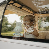 A child holding a Miffy Plush White & Vintage Stripes - 25cm bunny toy gazes out the window of a vehicle, with trees and greenery visible outside.