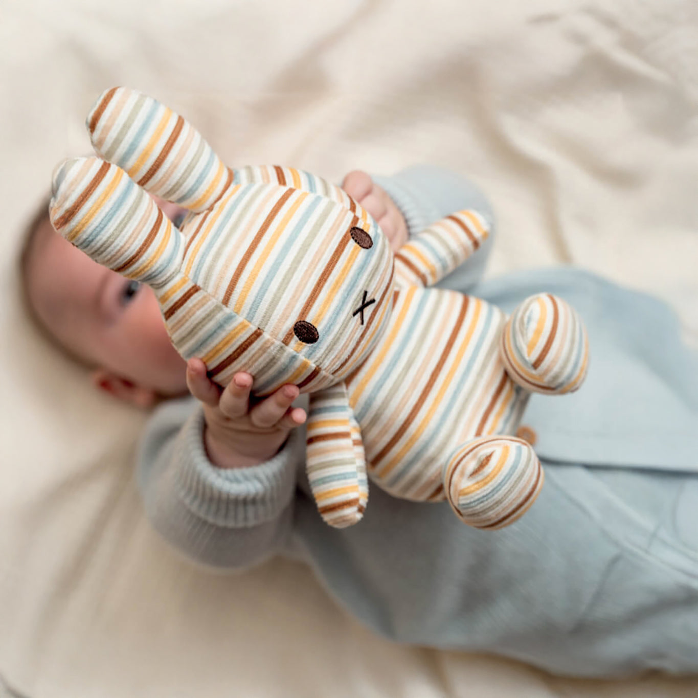 A baby lying down holds up a Miffy Plush Vintage Stripes All Over - 25cm toy, exuding timeless charm.