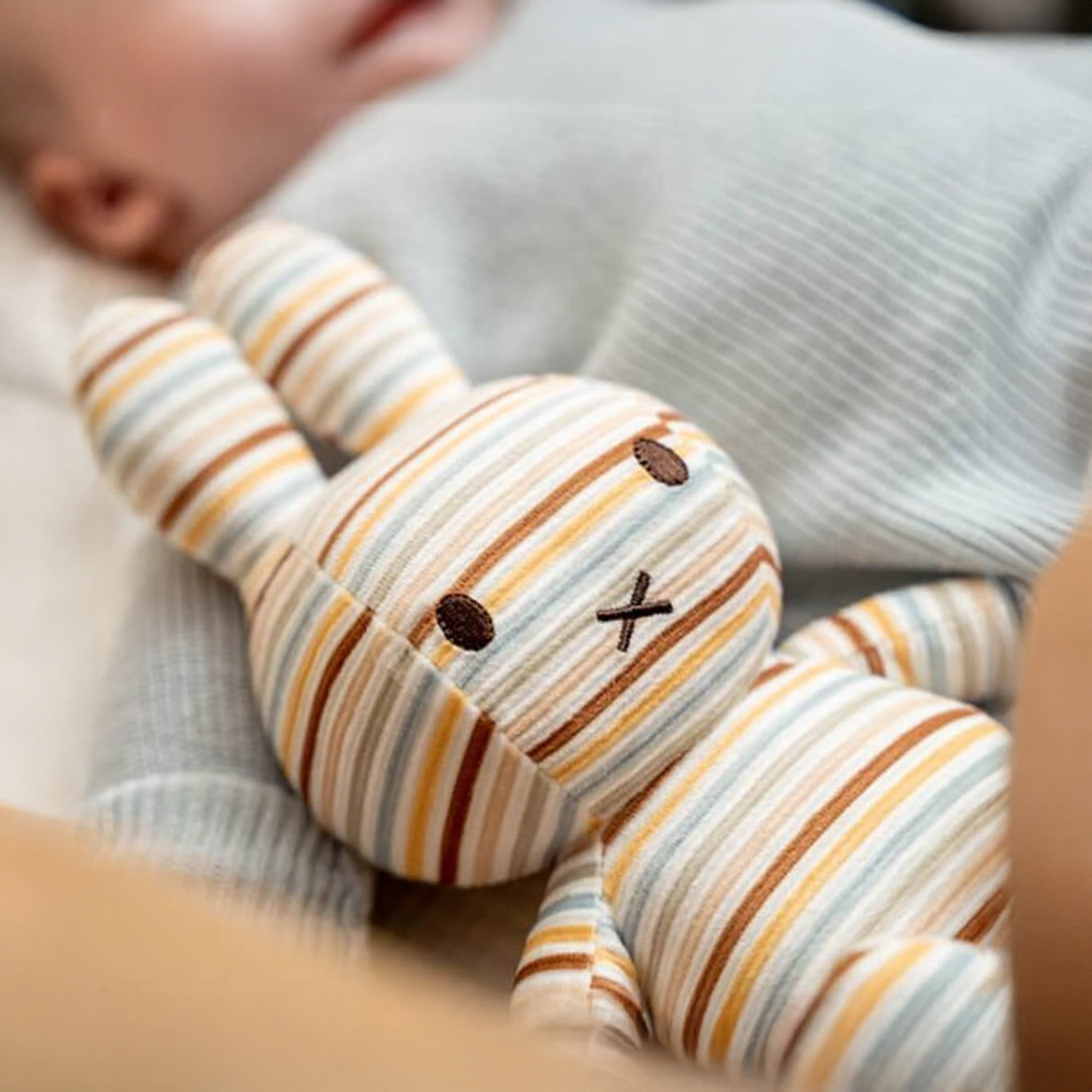 A Miffy Plush Vintage Stripes All Over - 25cm rests beside a sleeping baby.