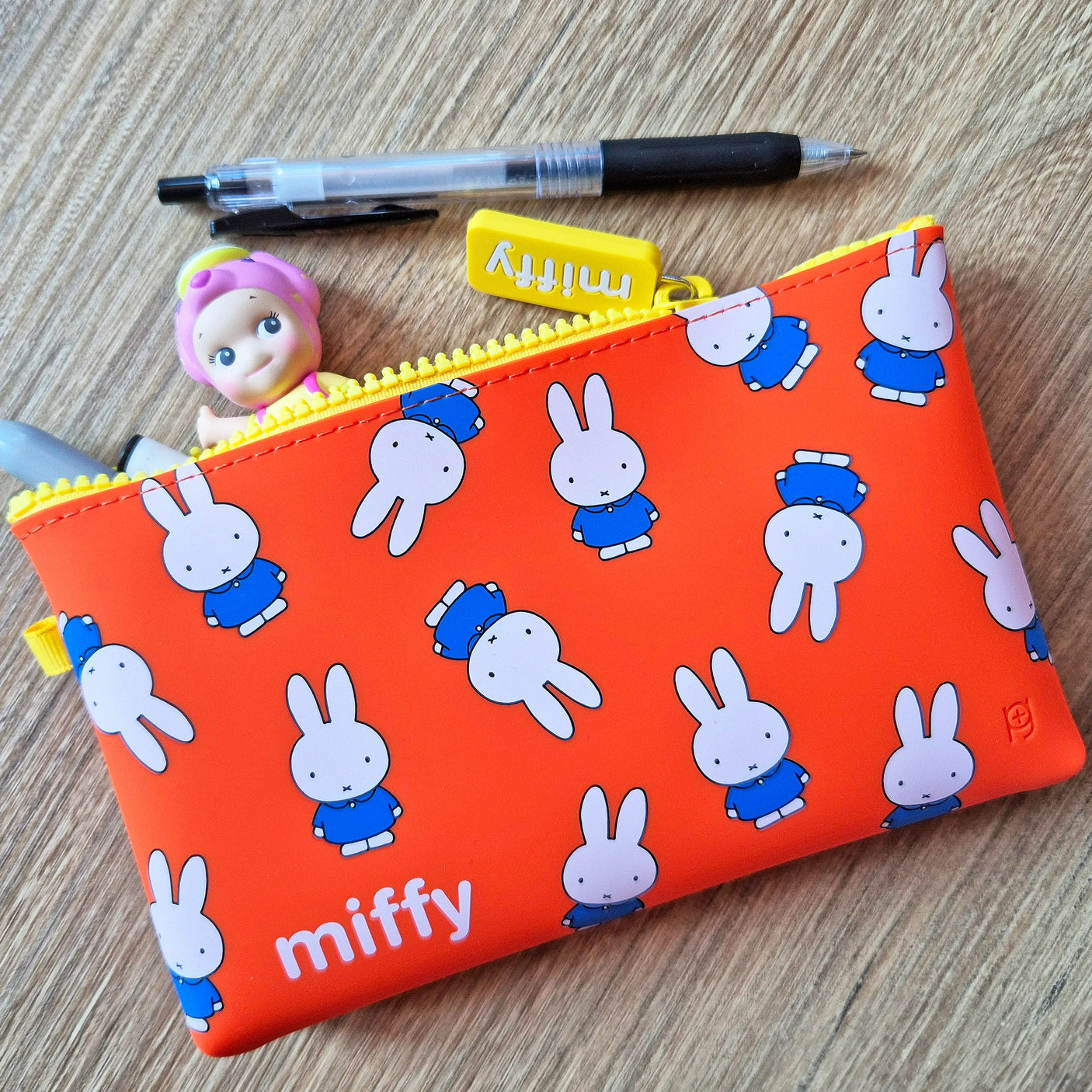 A Miffy Bunny Pattern Case 19cm, along with a pen and a small doll, rests on the wooden surface.