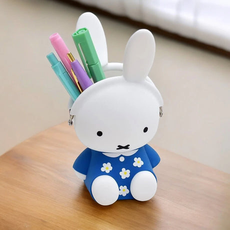 A Miffy Flower Dress Case Silicone, styled to look like a rabbit in a blue floral dress, holds colorful pens on a wooden table.