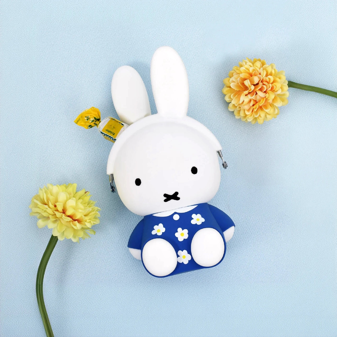 A Miffy Flower Dress Case Silicone, featuring a toy rabbit in a blue floral dress adorned with yellow tags, is charmingly displayed alongside two yellow flowers on a light blue backdrop.