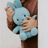 A child holds a Miffy Bunny Plush - Terry Light Blue 23cm, and stands against a white wall.