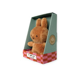 A Choc Beige Mini Miffy Plush from Miffy, resembling a snug brown rabbit toy, sits comfortably in a green and pink checkered gift box labeled "the label." The box highlights that it is made from "100% recycled plastic," making it an ideal Lucky Charm for any occasion.