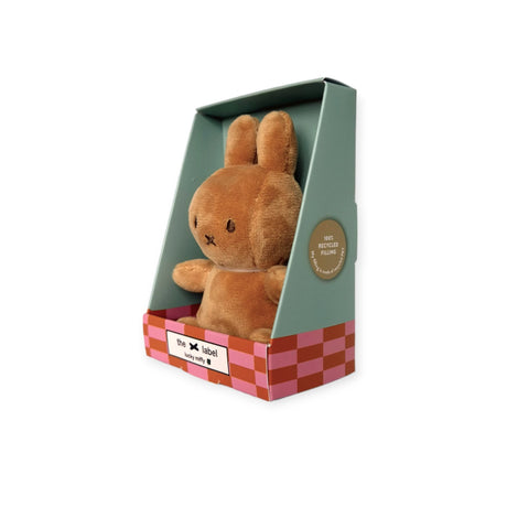 A Choc Beige Mini Miffy Plush from Miffy, resembling a snug brown rabbit toy, sits comfortably in a green and pink checkered gift box labeled "the label." The box highlights that it is made from "100% recycled plastic," making it an ideal Lucky Charm for any occasion.