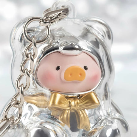 Clear bear-shaped keychain with pink pig figure wearing shiny silver hood and gold bowtie on blurred background