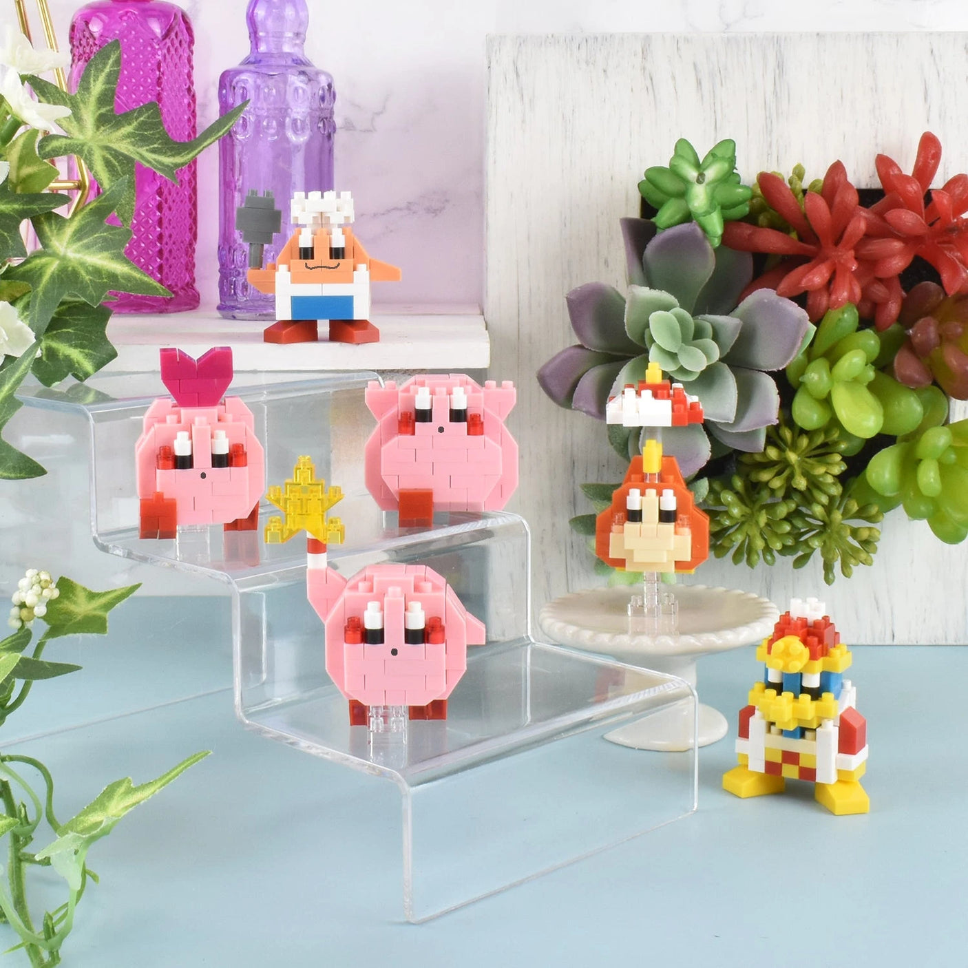 Toy figures in vibrant pixel-style, featuring characters like pink and orange Kirby figures from the Nanoblock Kirby Blind Bag - Vol. 2 series, are showcased on clear risers amidst plants and decorative accents.