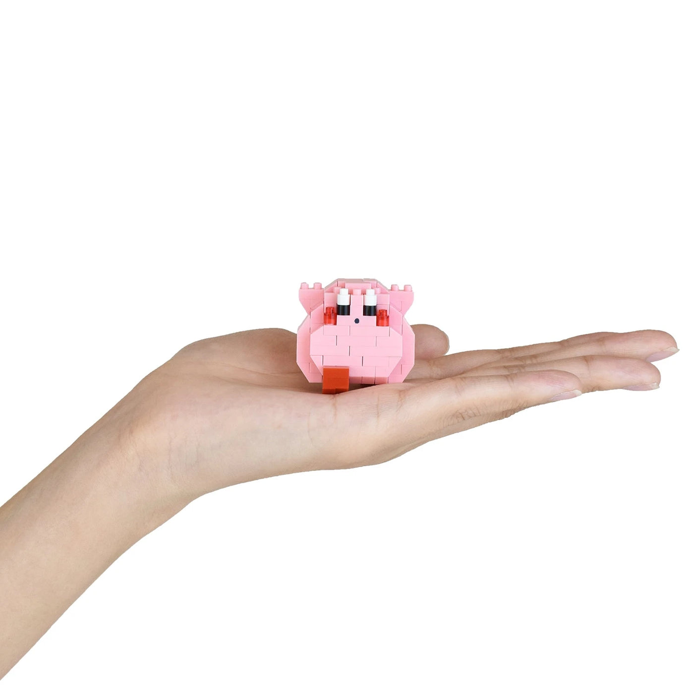 A hand holds a small pink toy resembling a pig from the Nanoblock Kirby Blind Bag - Vol. 2 series, featuring white and blue eyes, on a white background.