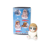 A small toy figurine of a cat wearing a brown helmet is placed next to a Mofusand Premium - Aquarium Cat Blind Box showcasing cartoon images of aquarium cats in various costumes.