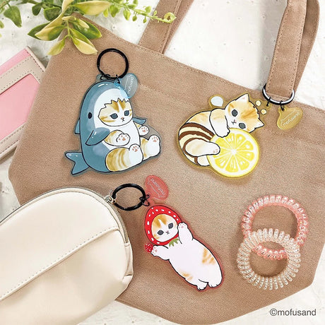 A beige tote bag with a Mofusand Shark Keychain, two cat-themed translucent acrylic keychains, a white pouch, pink wallet, pink hair ties, and a green plant in the corner.