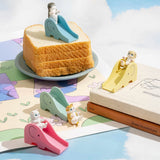 Toy cats on bright slides are whimsically arranged on bread and books, set against a sky-themed mat in the background, creating a playful scene reminiscent of the Mofusand Premium Blind Box - Cats on Slides collection.