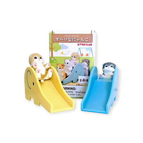 Mofusand Blind Box - Cats on Slides displays toy cats sliding in front of a colorful package featuring cartoon cats, complete with charming mini cat figures and a visible warning label at the bottom.