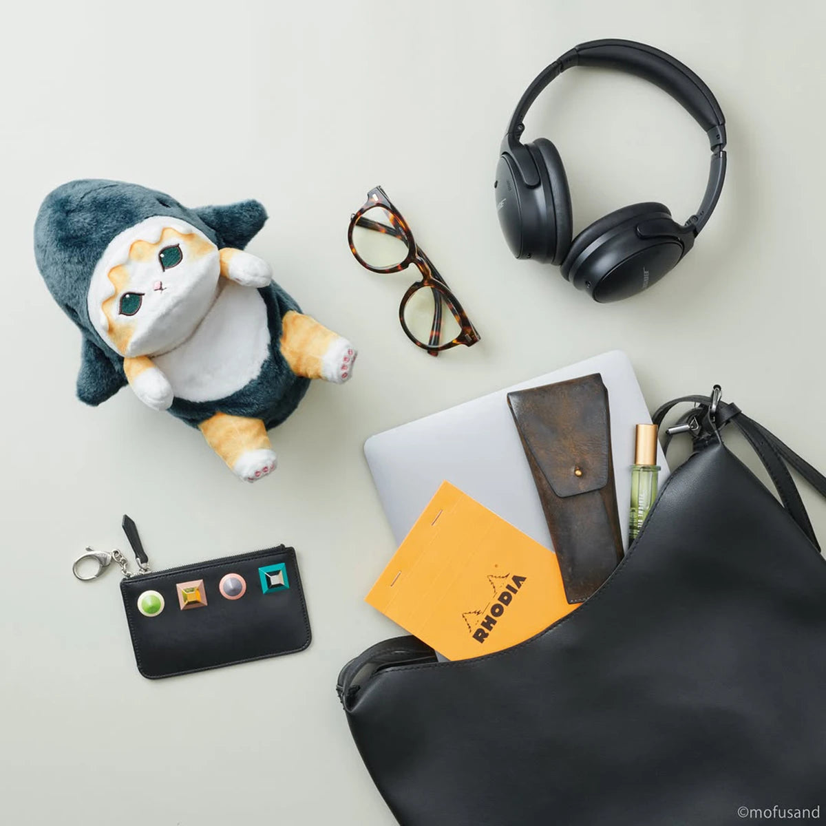 A Mofusand Posture Plush Buddy - Shark, headphones, eyeglasses, key pouch, laptop, notebook, pen case, and perfume bottle are neatly placed near a black shoulder bag on a light surface.