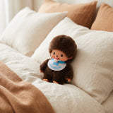Brown fur plush doll with blue bib and pacifier, sitting on bed