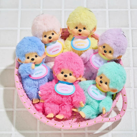 The Monchhichi Colours Bag Charm - Blue by Monchhichi features a plush doll with a round face, fluffy fur, and bib, perfect as a keychain or bag charm gift. Shown in a pink basket with pastel variants on a tiled surface.