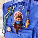 Brown plush Monchhichi keychain with blue scarf hanging from a blue backpack