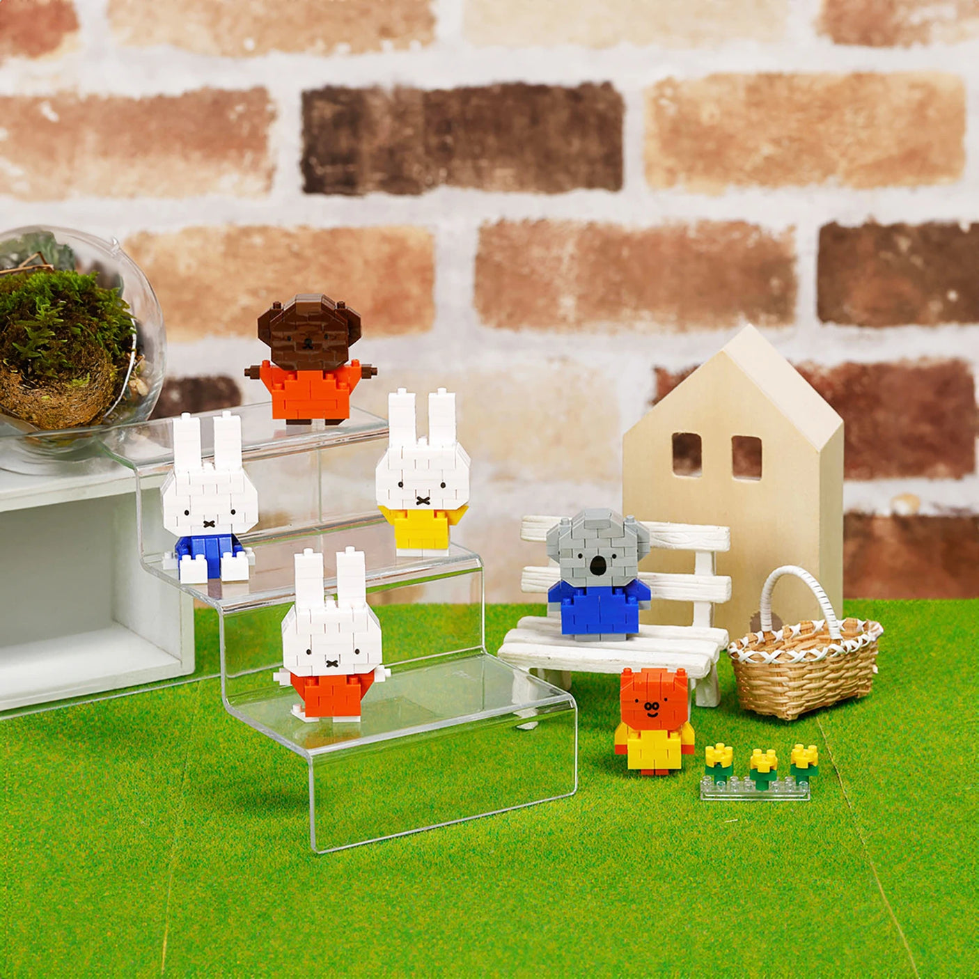 Animal-like toy figures, such as the Nanoblock Miffy Blind Bag - Vol. 3 figures, are showcased on transparent stands and a bench. A tiny house and basket are set in the background on a green surface, crafting a delightful scene reminiscent of a Blind Bag surprise by Nanoblock.